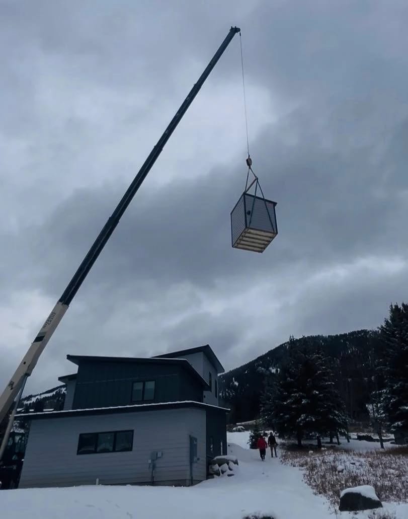 A crane lifting a custom sauna into place beside a house on a snowy day.