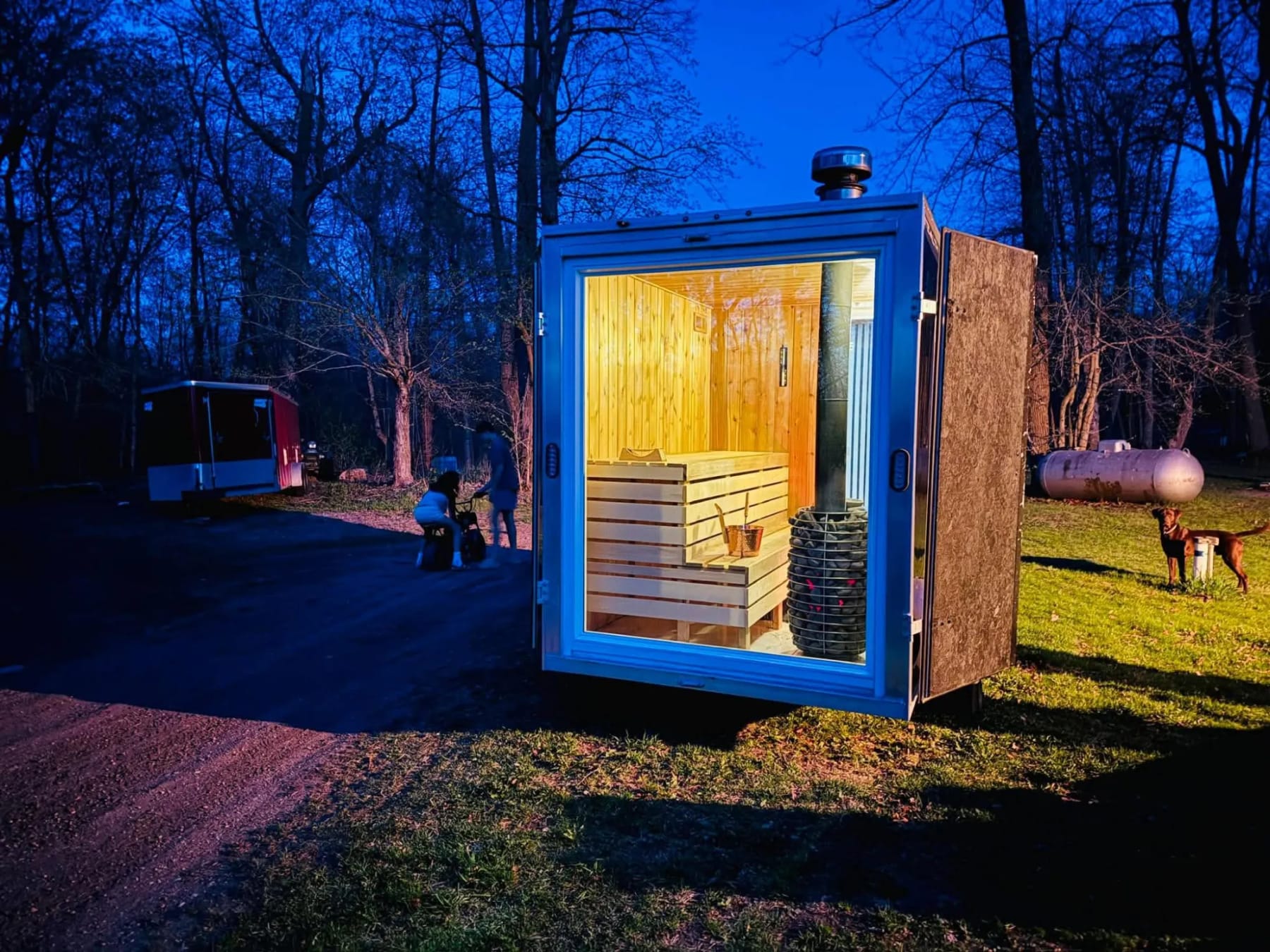 Mobile sauna lit up at dusk, its benches and wood stove showing through the glass front.
