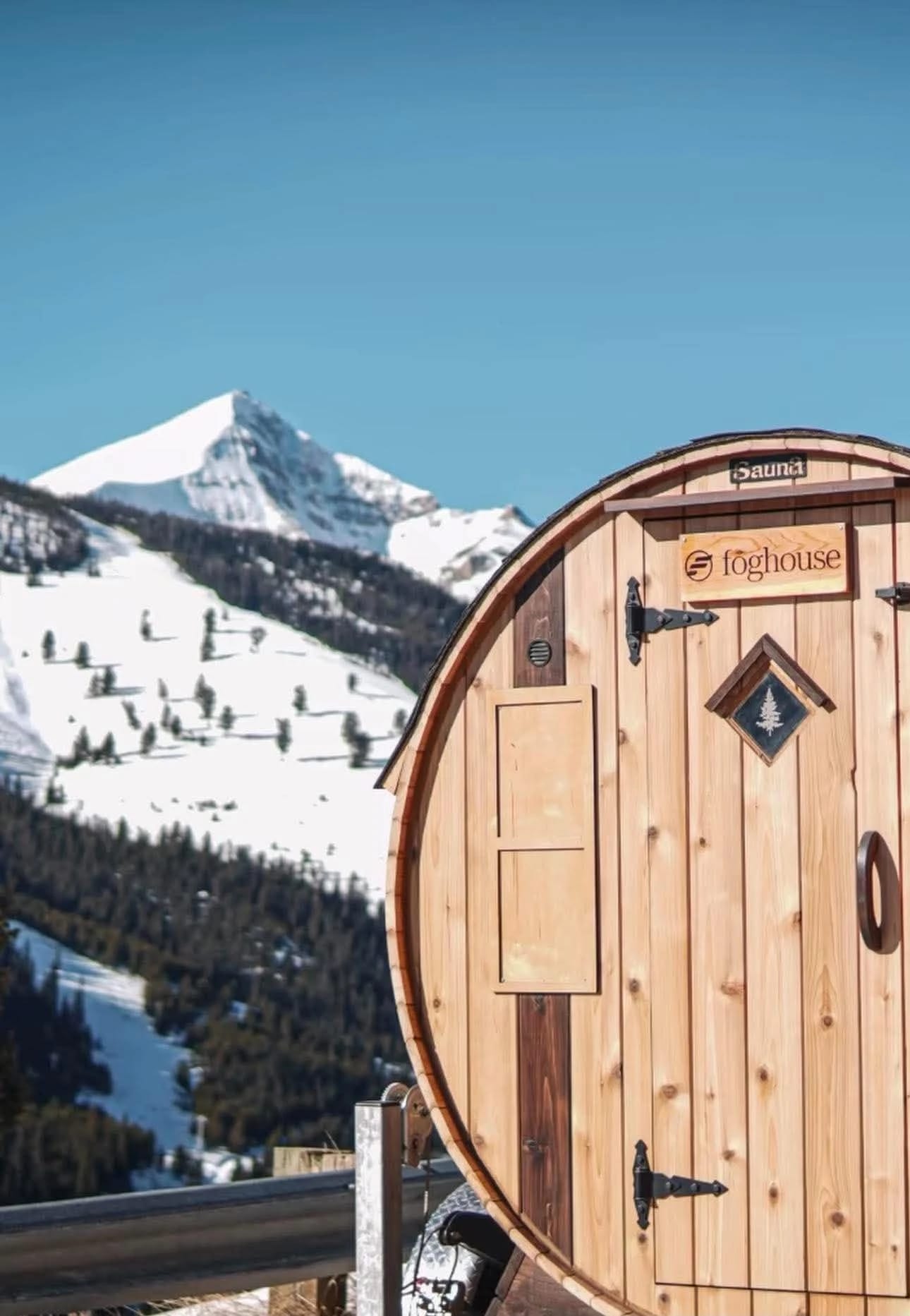 Cedar barrel sauna in front of a snow-covered mountain on a clear day.